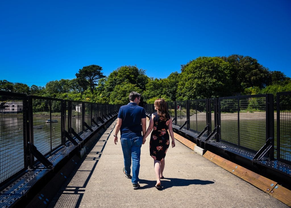 a man and woman holding hands walking on a bridge over water