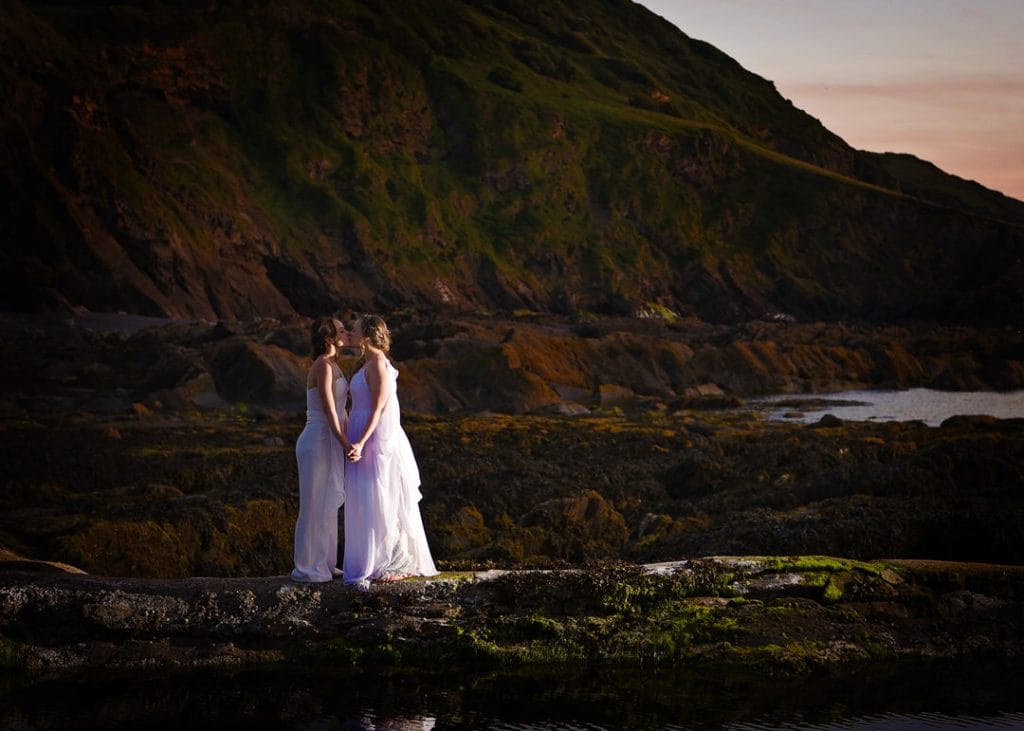 Two people in white dresses stand hand in hand on a rocky shoreline at sunset. A large, green hill serves as a backdrop, with a calm body of water nearby. The scene conveys a sense of tranquility and connection.