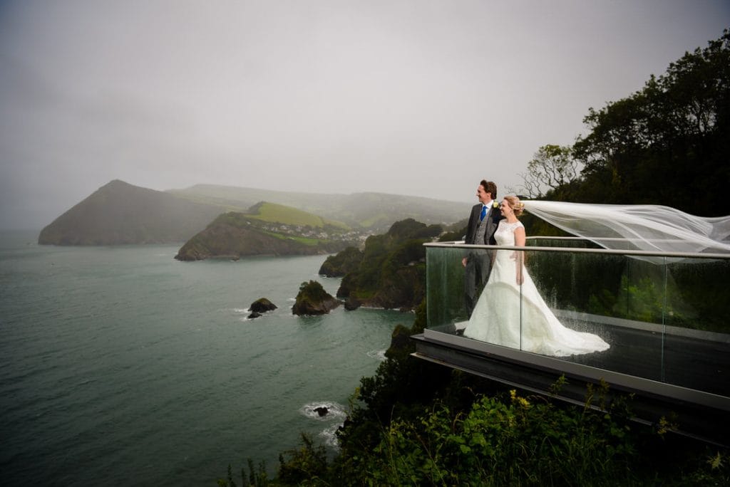 A bride and groom stand on a glass balcony overlooking a foggy, rugged coastline. The brides veil flows in the wind. The sea is a calm turquoise, with cliffs and lush greenery in the background under an overcast sky.