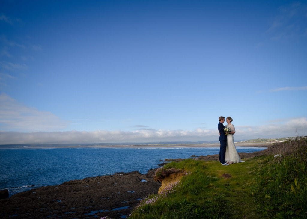 A bride in a white dress and a groom in a blue suit stand together on a grassy cliff overlooking a vast, calm ocean. The sky is clear with a few scattered clouds, and distant land is visible on the horizon.