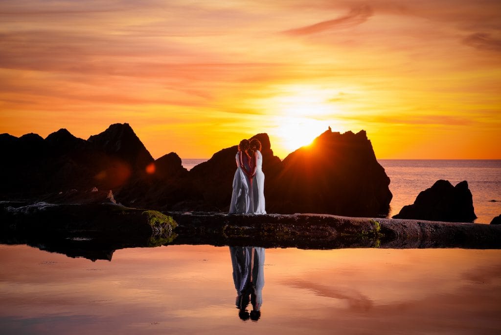 a couple of people standing on a rock in front of water with a sunset